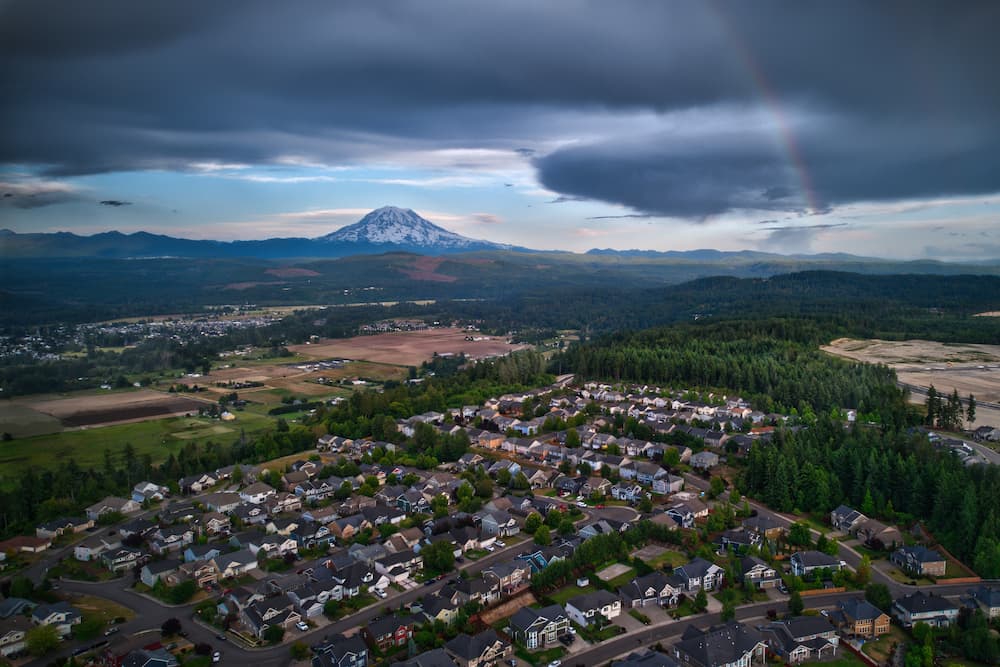 Drone photo of a faint rainbow dimly illuminating a thin section of cloudy skies surrounding Mt. Rainier, Washington.
